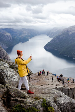 Smiling blonde woman hiker in yellow raincoat stays on the top of the Preikestolen mountain (Preacher's Pulpit or Pulpit Rock) with a lot of tourists and shoots photos by phone on Lysefjord background