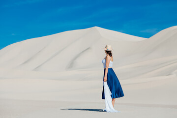 Woman walking alone in the desert dunes landscape and looking at horizon line. Girl dressed in blue skirt, white top, white scarf and hat sitting in Dumont dunes, California in Mojave Desert.