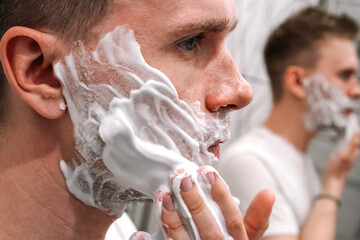 A young man puts shaving foam on his cheeks