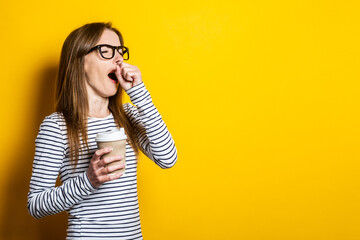 Portrait of a yawning young woman holding a paper cup with a coffee on a yellow background