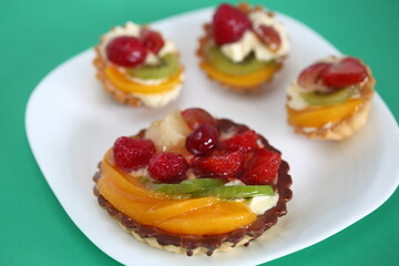 fruit cakes with strawberries, kiwi fruits, raspberries, peach and cream on white plate close up isolated on light green background
