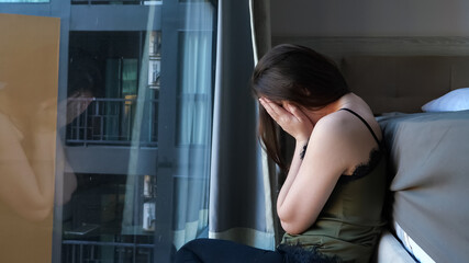 upset sleepy woman with long loose hair sits near large bed and window in comfortable hotel room in early morning