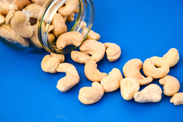 close up heap of roasted cashew nuts spilling out of glass jar scattered on blue background, seed...