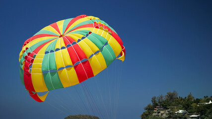 wonderful red green yellow parachute moves under clear blue sky against green hilltop with small buildings silhouettes closeup