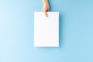 Woman’s hand holding empty white paper sheet on blue background. Close up