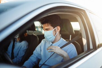 Young boy taxi driver fasten the seat belt wearing sterile medical mask. A man in the car behind the steering wheel works during coronavirus pandemic. Social distance and health safety concept.