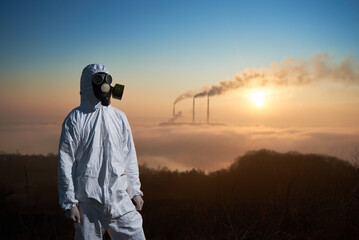 Portrait of a scientist wearing white protective uniform and gas mask standing on a hill, smoking pipes of a thermal power plant are on background. Concept of environmental pollution.