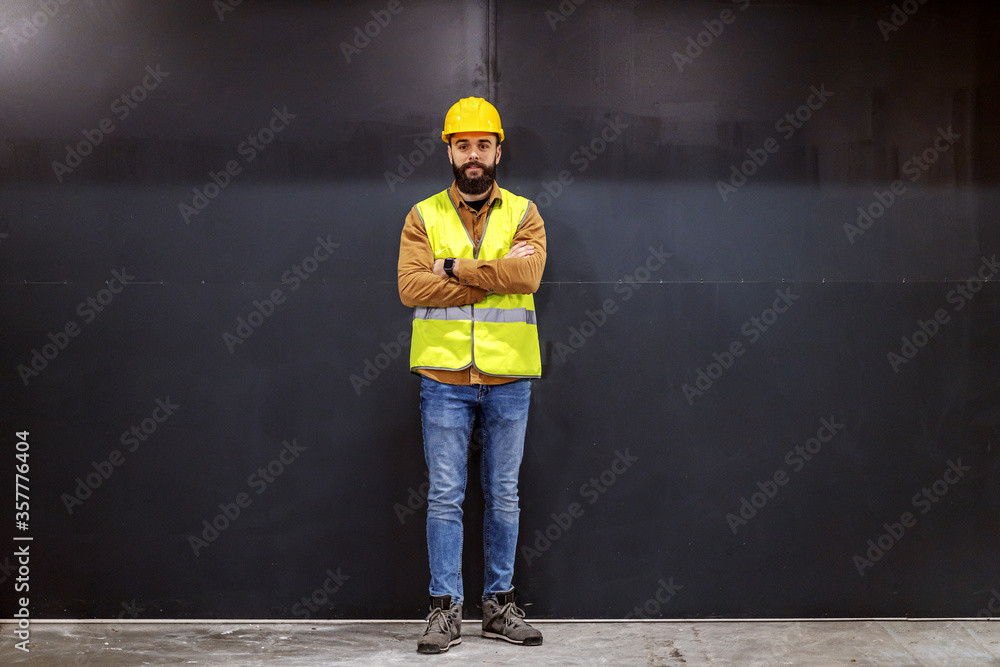 Wall mural young smiling attractive bearded worker in vest with helmet on head standing in front of black door 