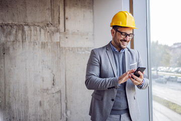 Smiling architect in suit and with helmet on head standing in building in construction process next to window and typing message on smart phone.