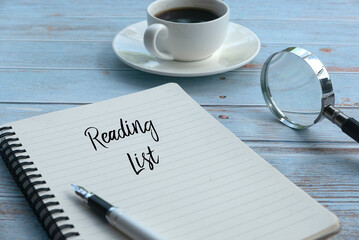 Selective focus of a cup of coffee,magnifying glass,pen and notebook written with Reading List on wooden background.