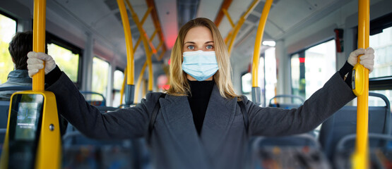 Fototapeta premium Young blonde in medical mask looking at camera while holding handrail while standing in bus lounge.