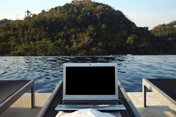 Fototapeta premium woman with laptop resting alone in lounge chair at hotel pool against forestry hill close back view