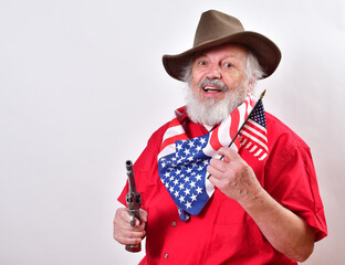Smiling old cowboy with a pistol in one hand and the American flag in the other..Rancher is celebrating the 4th of July with a patriotic bandana, pistol and American flag.