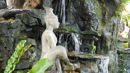 beautiful white stone meditating Buddha statue located near artificial waterfall on rocks with green grass and small temple model
