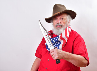 Bearded man wearing a patriotic bandana and a floppy western hat is showing off his giant hunting knfe..Cowvoy using his giant bowie knife to show off.. - obrazy, fototapety, plakaty