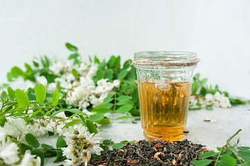 acacia flowers broth in glassware cup on a white cement background with lush flowering white acacia tree Branch , home self-treatment, useful properties of plants