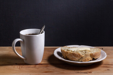 A white Cup with a spoon and coffee, on a wooden Board, a white plate with a slice of homemade bread and butter.