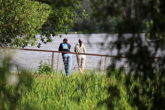 Love couple standing on a river coast in a park, view through tree leaves. Summer leisure, romantic date on a nature
