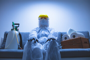 Person with toilet paper stock and protective antiviral mask, suit, helmet and chemical decontamination sprayer bottle in home isolation.