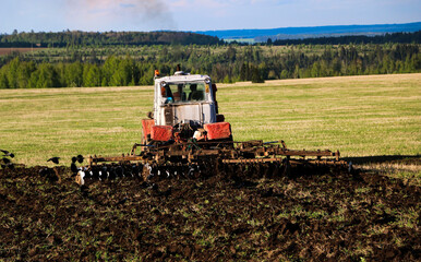 Fototapeta premium tractor plows and harrows land in large field on sunny spring day. preparing soil for planting crops, plowing soil with tractor with disk plow.