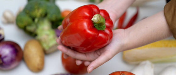Hands holding red bell pepper while standing at table with other fresh farm vegetables
