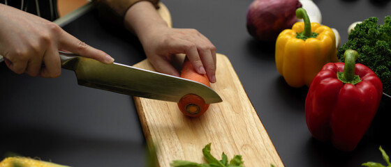 Female cook slicing fresh carrot on wooden chopping block at dark modern kitchen