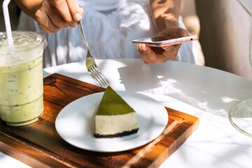 A girl is sitting in cafe with matcha green tea latte and cake on table. Female hands holding smartphone while eating a piece of green tea cheese cake on white table