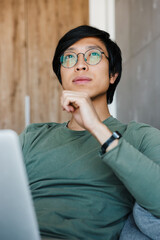 Image of thinking asian man working with laptop while sitting