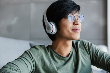 Image of joyful asian man using wireless headphones and smiling