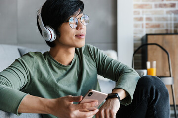 Image of smiling asian man using cellphone and wireless headphones