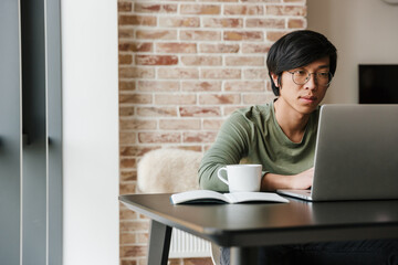Image of serious asian man working with laptop and drinking coffee