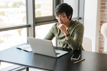 Image of serious asian man working with laptop while sitting at table