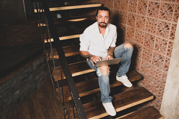 young handsome man working at home and siting on stairs with laptop on room background