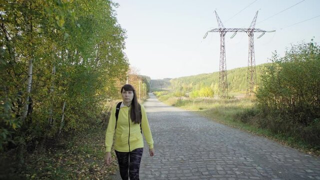 Young brunette woman in yellow sports jacket walks along the edge of cobblestone road through forest next to power line. Female hiker with backpack steps along the clearing on an autumn warm day. 