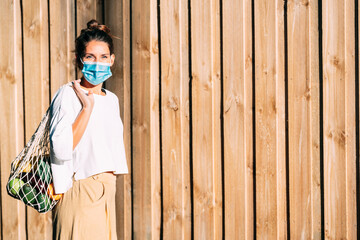 A green eyes woman with a face mask and a reusable mesh shopping bag full of fruits and vegetables