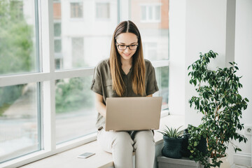 Beautiful young adult positive girl uses a laptop at home by the window or is she in open space.