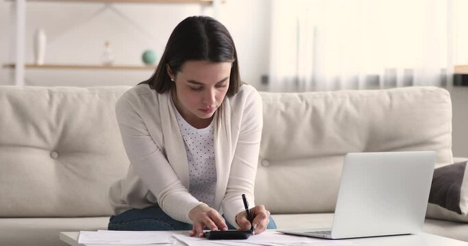 Focused serious smart young 20s woman sitting on sofa, calculating monthly budget, managing incomes and outcomes money cashflow, checking bills or rental payment, accounting taxes alone at home.