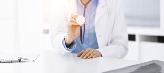 Unknown woman-doctor in blue blouse is holding stethoscope head while sitting at the desk in sunny clinic, close-up. Medicine concept