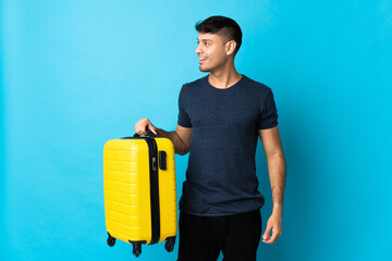 Young Colombian man isolated on blue background in vacation with travel suitcase