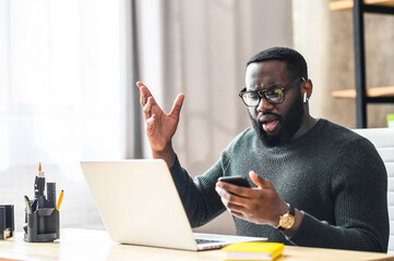 Confused African-American guy looks at phone screen while sitting at the office desk with a laptop,...