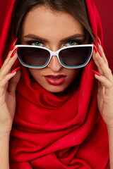 Portrait of classy brunette young woman with luxurious make-up in white sunglasses and red headscarf on red background. woman look at the camera