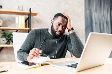 Frustrated sad black guy is watching at laptop screen and holding head with his hand. He read bad...