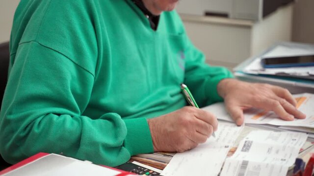 Working at home in coronavirus quarantine, paperwork in distance. Senior man working at the table in apartment room, old doctor writing down something with pen on sheet paper, reading and making notes