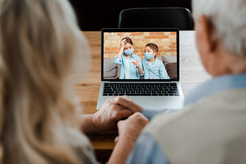 grandparents holding hands and having video chat with grandchildren in medical masks holding thermometer during self isolation