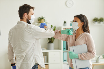 Waist up portrait of two colleagues wearing face masks bumping elbows while greeting each other at...