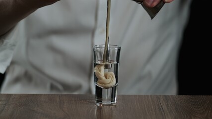 The bartender pours irish cream from jigger to cocktail glass with sambuca. Close up