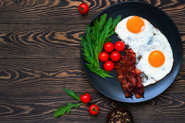 Homemade breakfast with fried eggs, bacon, cherry tomatoes and arugula  served on the dark wooden table. Top view. Closeup.