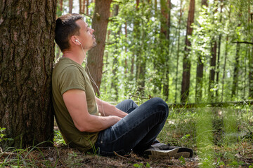 Obraz premium a man in a forest lean against a tree, with headphones listening to music. rest at nature. in the background blurred silhouettes of trees. digital detox. Mental health. Positive emotion.