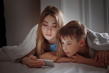 A young girl shows her little brother a video on her phone while lying on a bed at night. Brother and sister are watching a video on their phone.