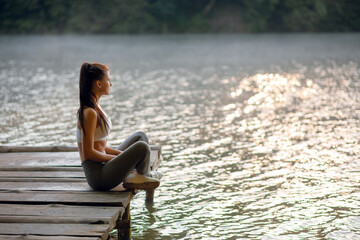 Beautiful sporty woman in stylish sportswear sitting on the wooden pier on the lake and enjoying beautiful nature.